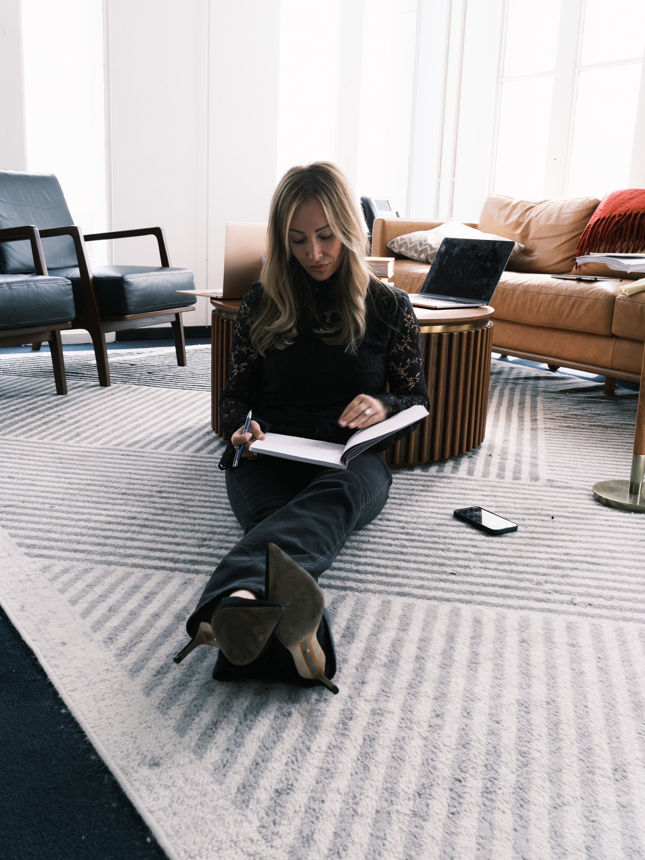 A woman sitting on a striped rug in a modern office or living room, writing in a notebook with a pen. She has blonde hair and is dressed in black, with a backpack and several laptops behind her, along with a smartphone on the floor.