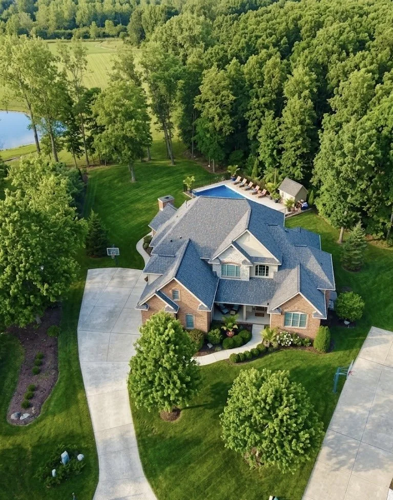 Aerial view of a large suburban house with a gray roof, surrounded by lush green lawn, mature trees, a backyard with a swimming pool and lounge chairs, and concrete driveways.
