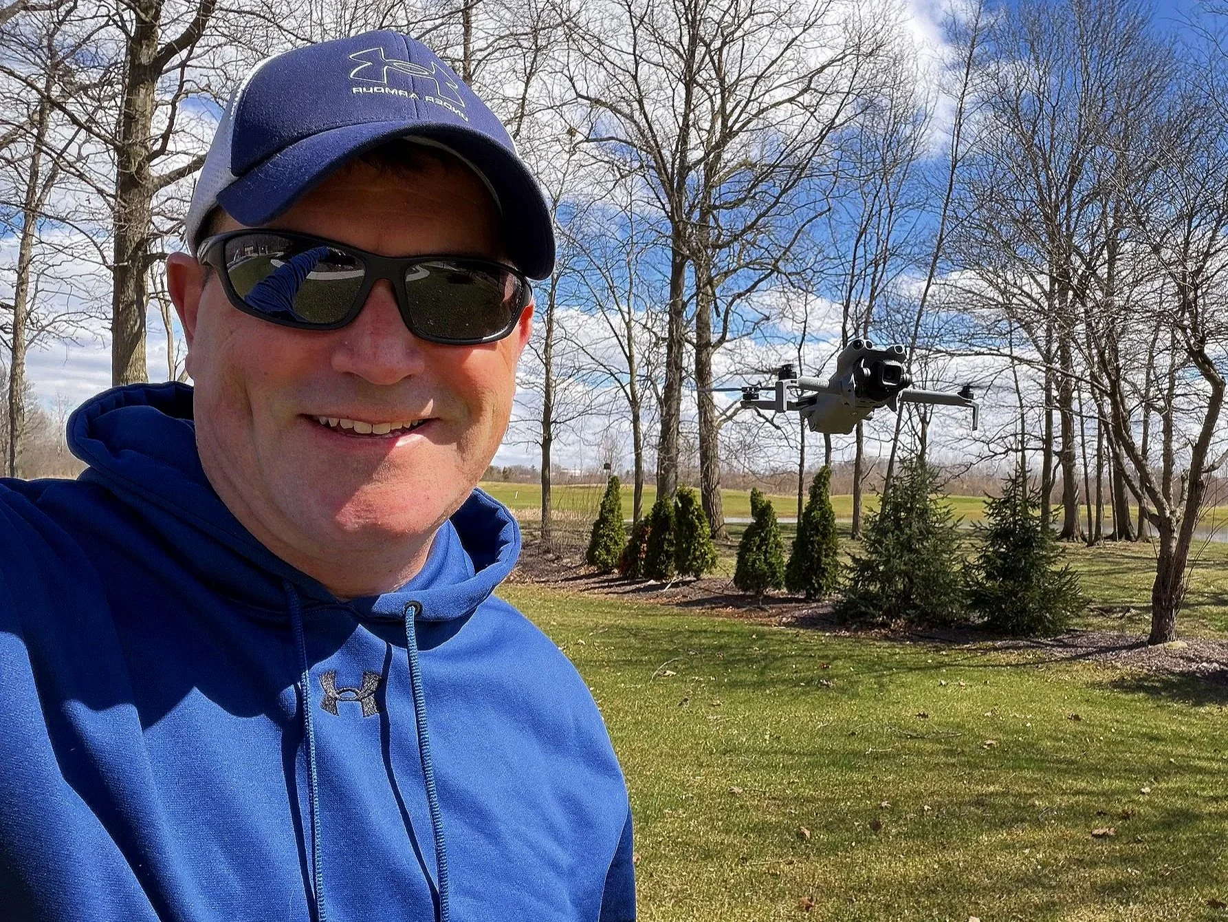 A man in sunglasses and a blue cap and jacket taking a selfie in a park with leafless trees and a drone flying in the background.