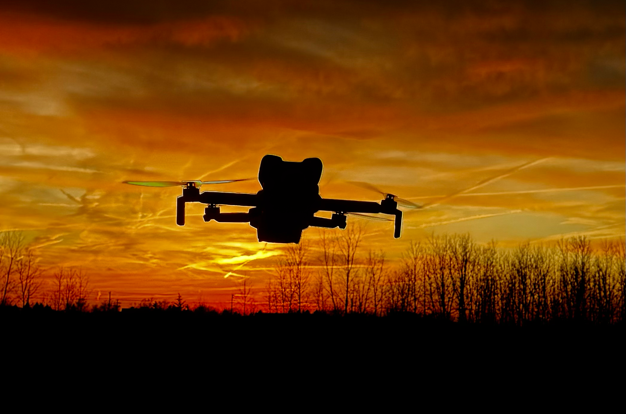 Silhouette of a drone flying against a vibrant sunset sky with orange and yellow clouds, with trees lining the horizon.