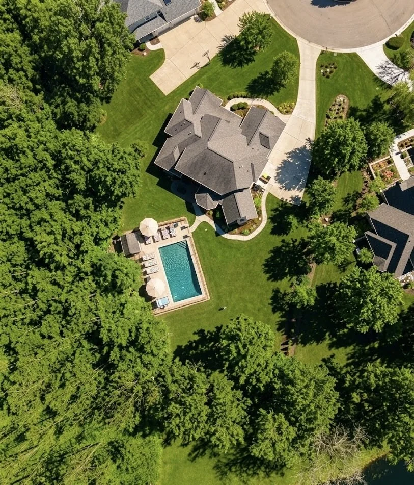 Aerial view of a residential backyard showing a house with a gray roof, a swimming pool with lounge chairs and umbrellas, green lawns, mature trees, and nearby houses.