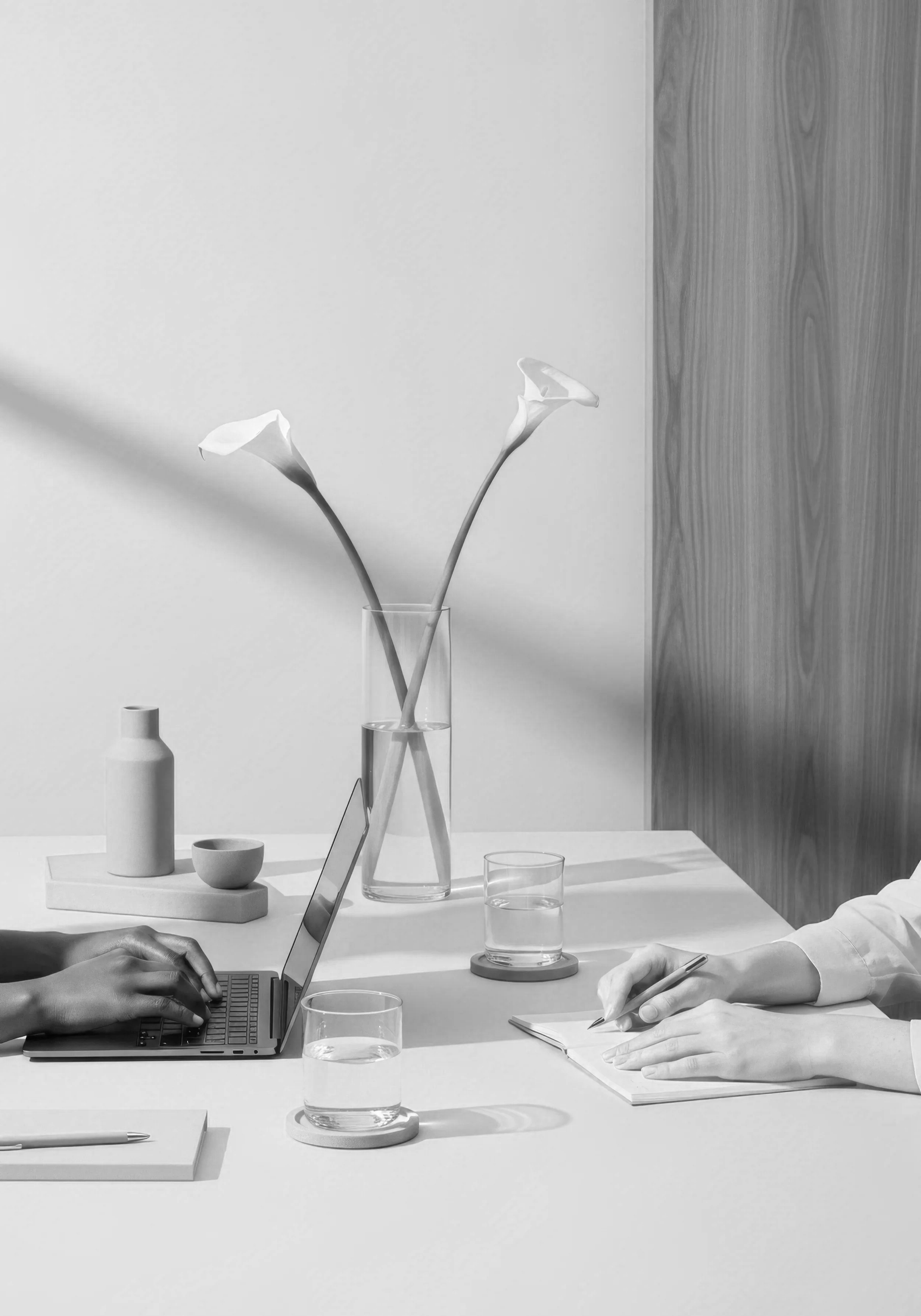 A minimalist workspace with two glasses of water, a laptop, a notebook, and two hands, one typing on the laptop and the other writing in the notebook. A tall glass vase with two calla lilies is on the desk, alongside small decorative items and a wooden panel background.