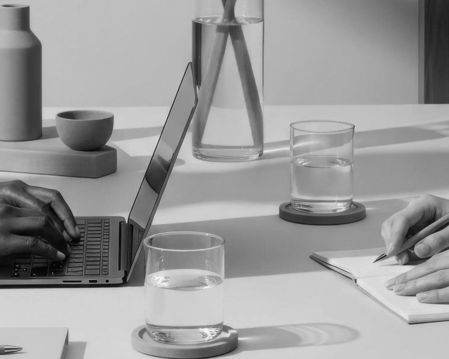 Black and white photo of a workspace with a person typing on a laptop and another writing in a notebook, with glasses of water, a vase, a bowl, and other objects on the desk.
