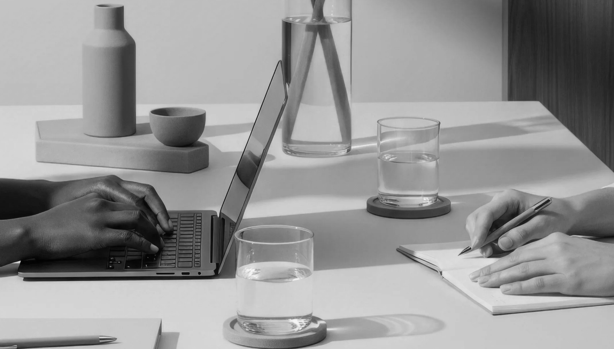 Two people working at a white table, one typing on a laptop and the other writing in a notebook, with glasses of water and minimalistic decor.