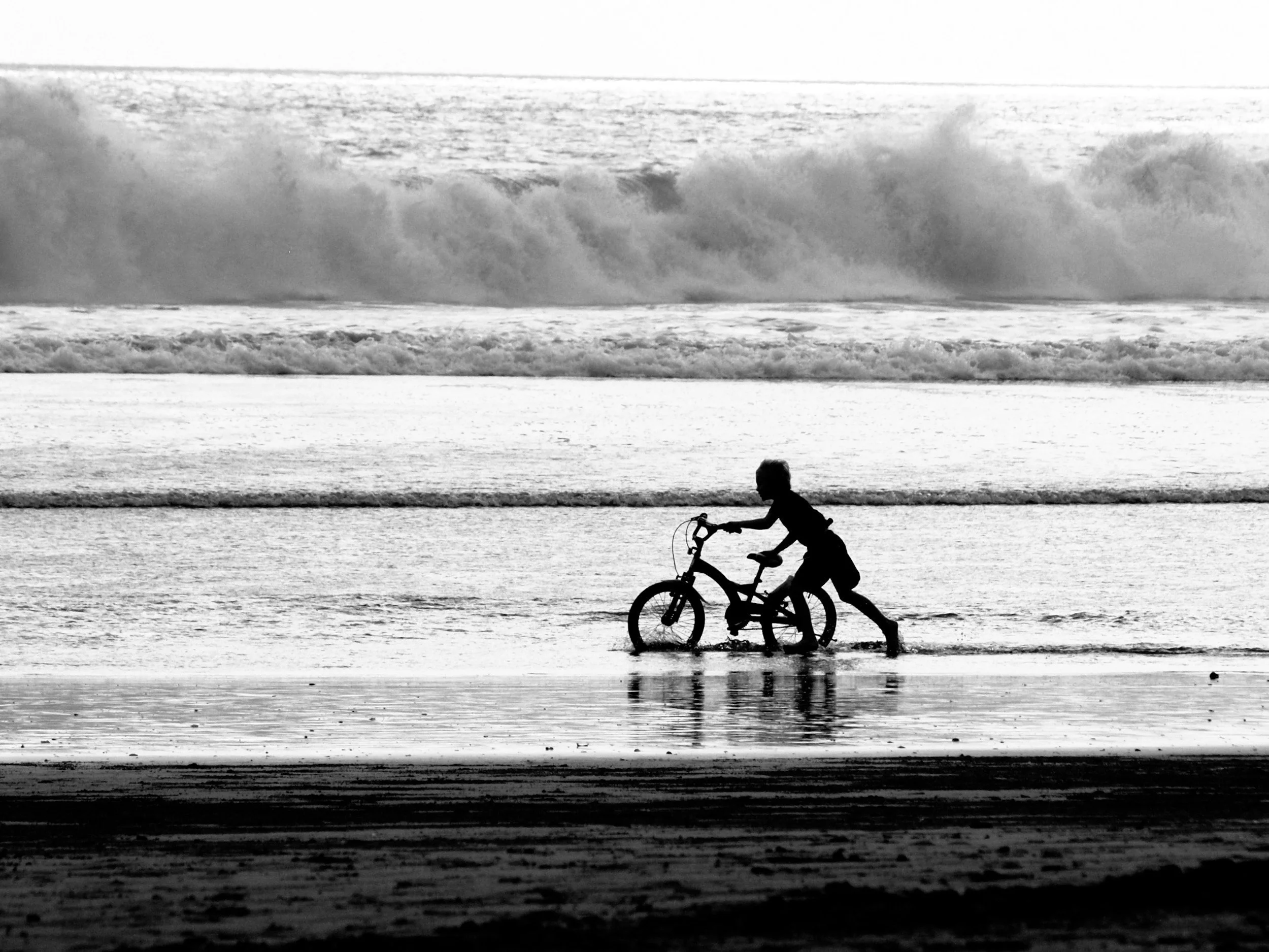 Child pushing a bicycle in shallow ocean water at the beach, with large waves in the background, in black and white.