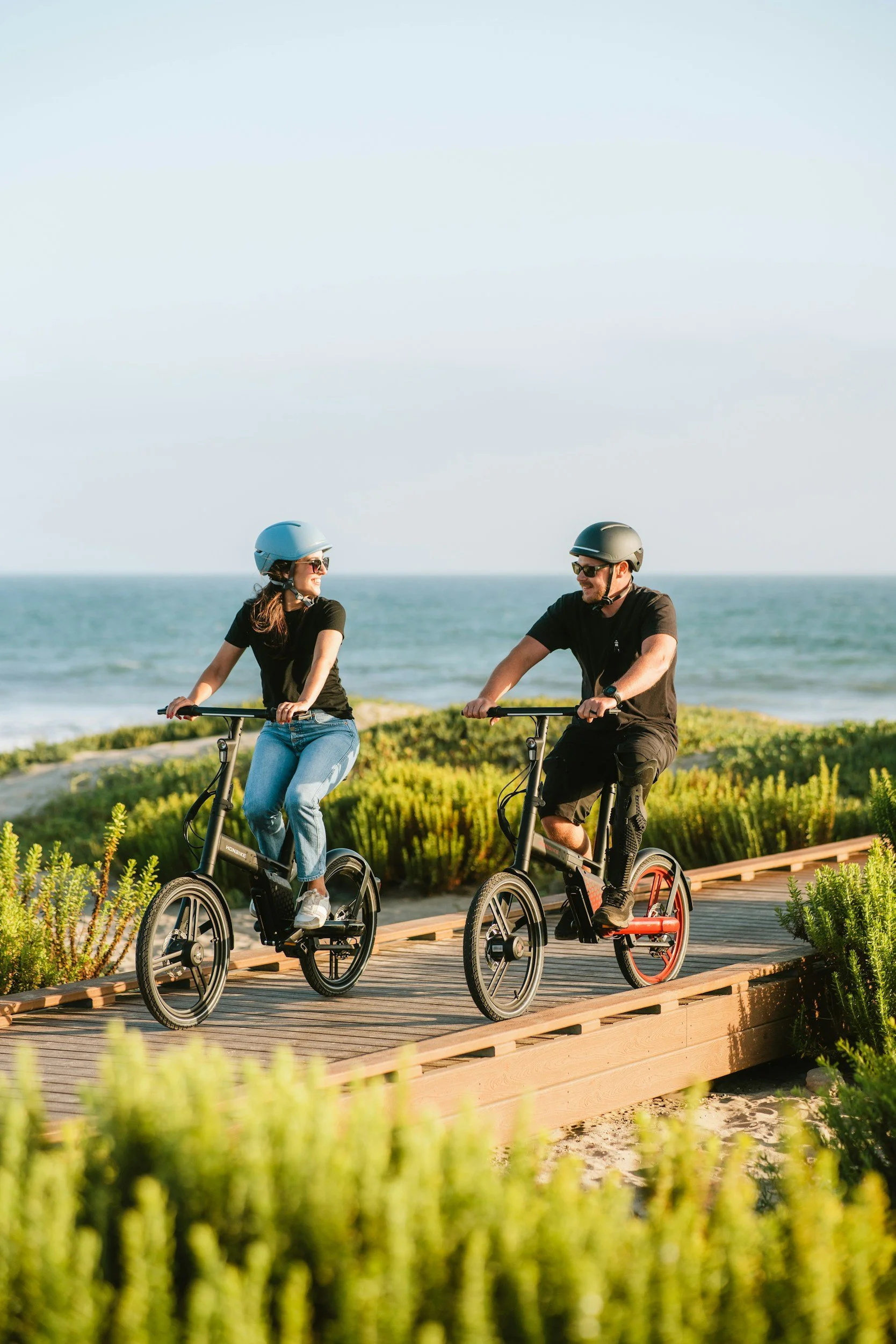 A man and woman riding electric bikes on a wooden path near the beach, wearing helmets and sunglasses, enjoying a sunny day with the ocean in the background.