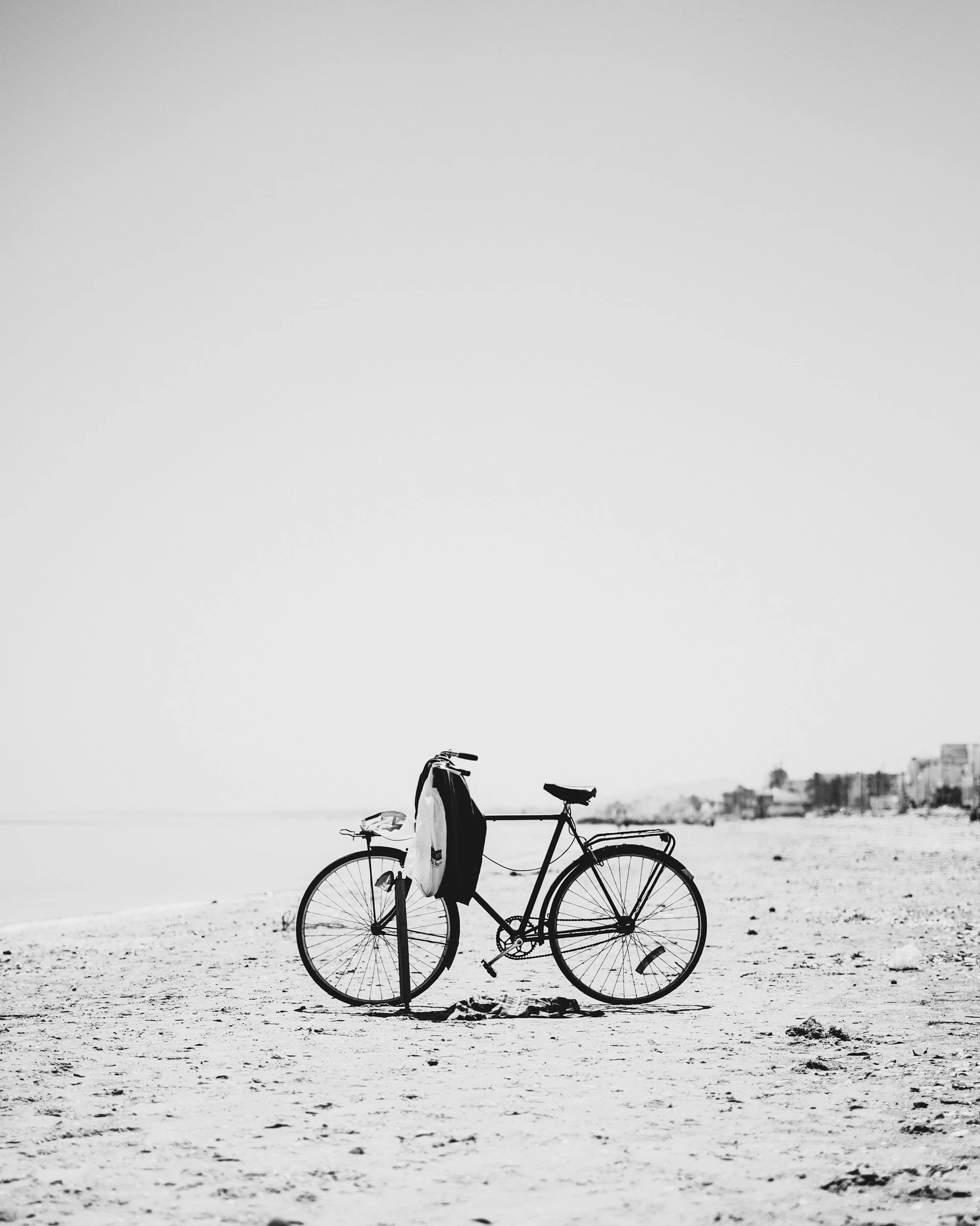 A black bicycle with a bag and jacket hanging from the handlebars, parked on a sandy beach near the shoreline under a clear sky.