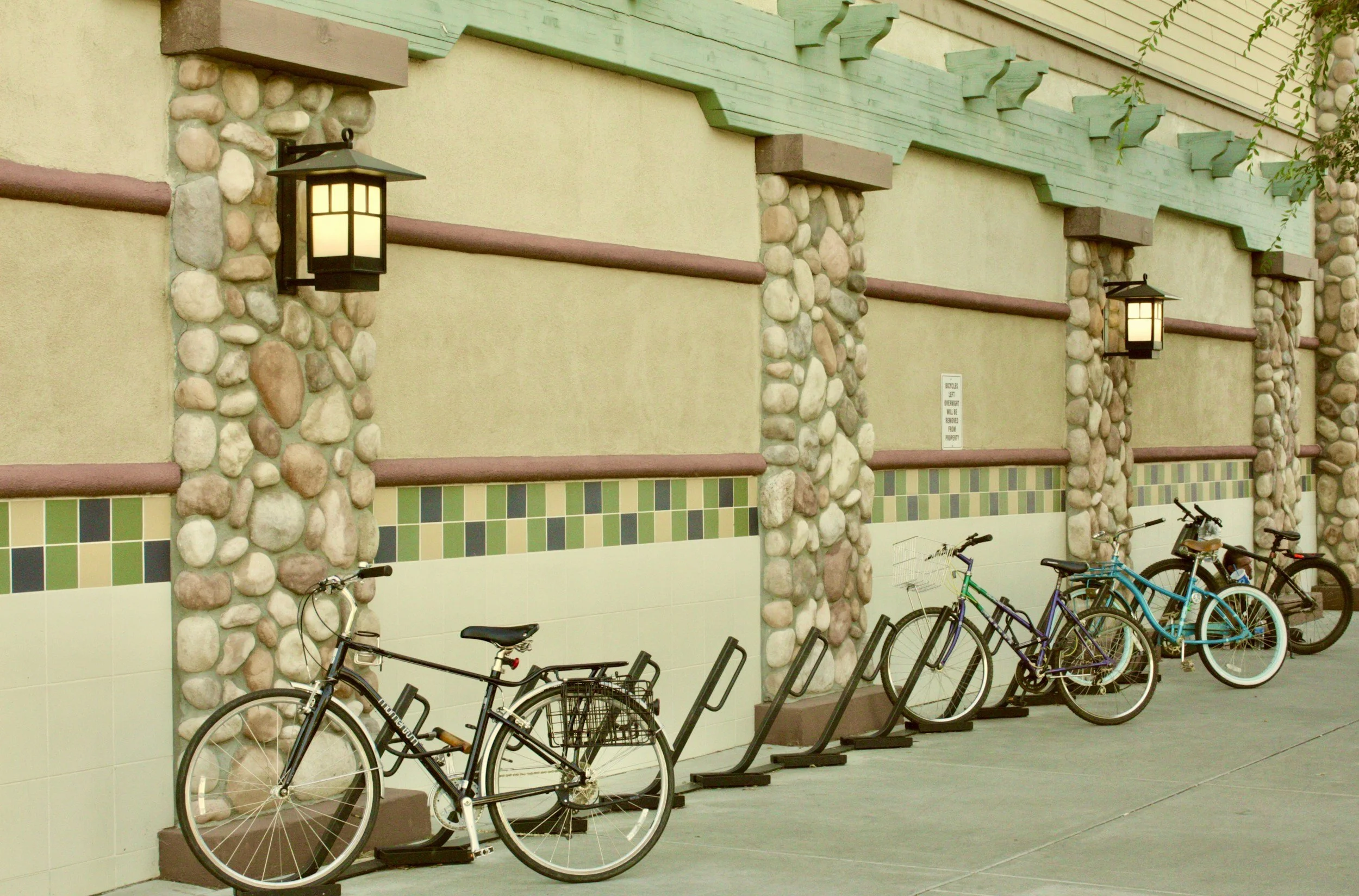 Row of bicycles parked at a bike rack against a decorative wall with stone and tile accents, illuminated by wall-mounted lanterns.