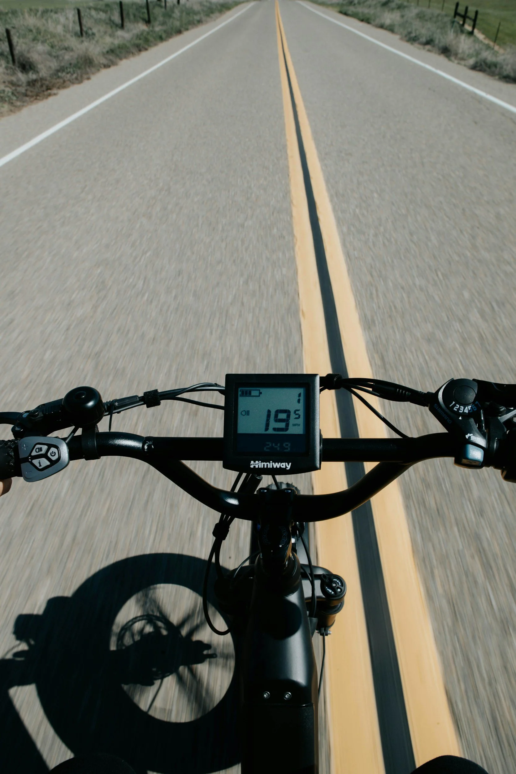 View from a bicycle on a paved two-lane road with yellow center lines, open countryside on both sides, digital display on handlebars showing 19.5 mph, 24.9 miles, and battery level.