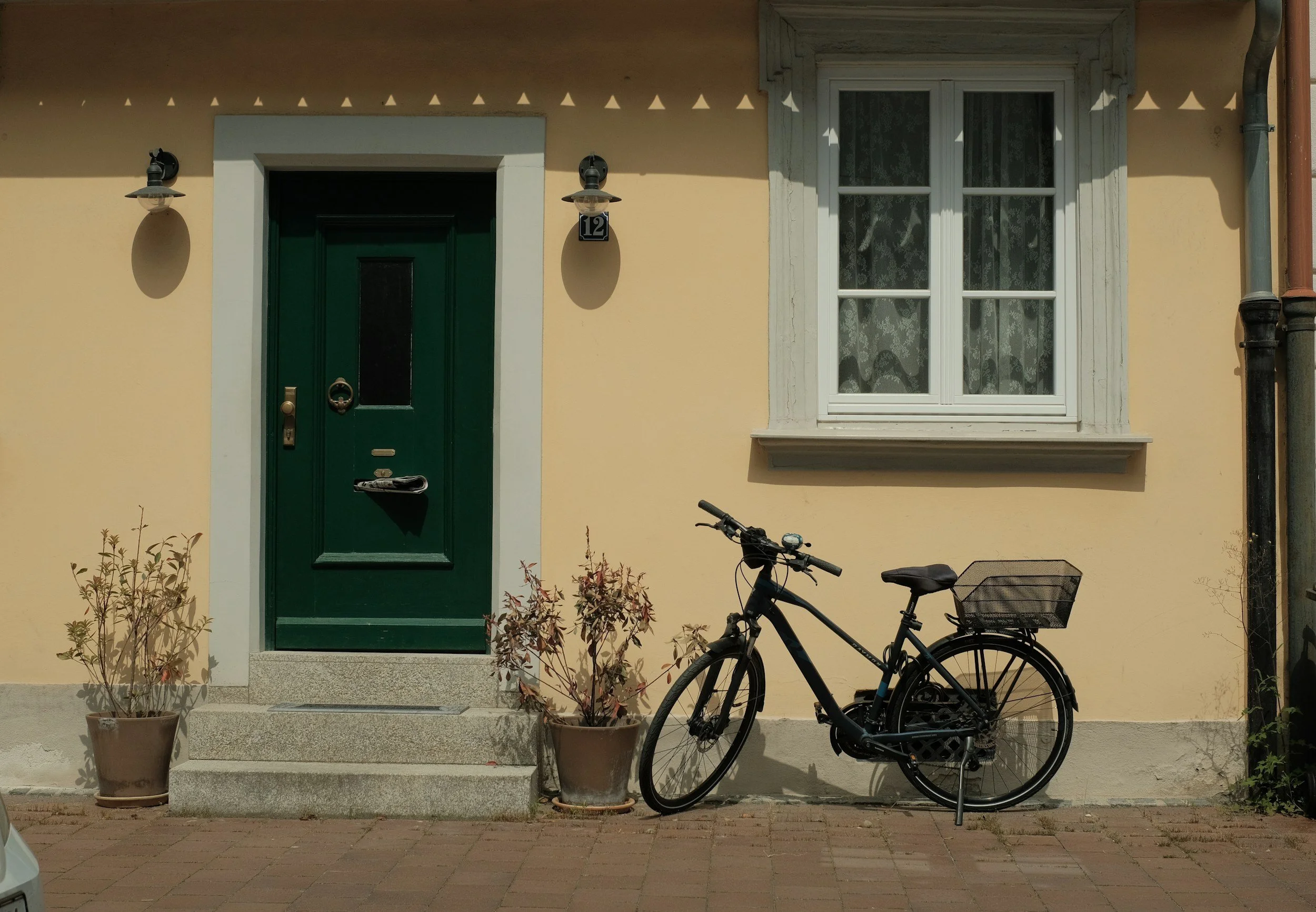 A house entrance with yellow walls, a green door, and two potted plants on the steps. A black bicycle with a basket is parked to the right of the door, and there is a window with white framing and lace curtains to the right of the door.