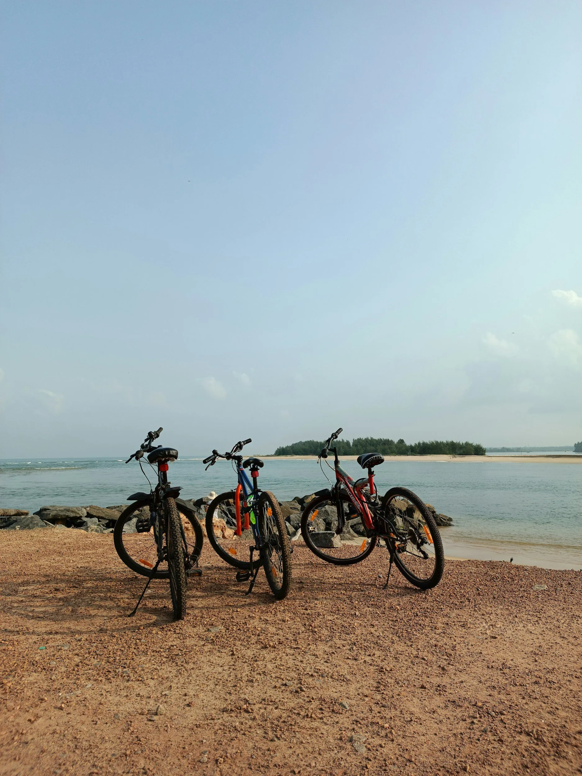 Three bicycles parked on sandy beach near water with rocks and a small island in the background during daytime.