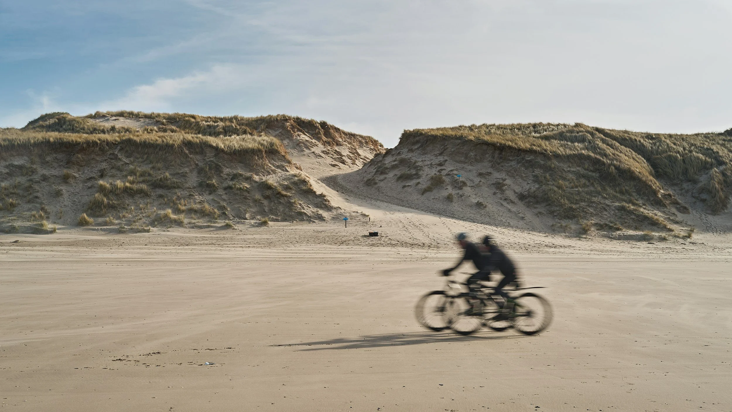 Two people riding bikes on a sandy beach with dunes in the background and a partly cloudy sky.