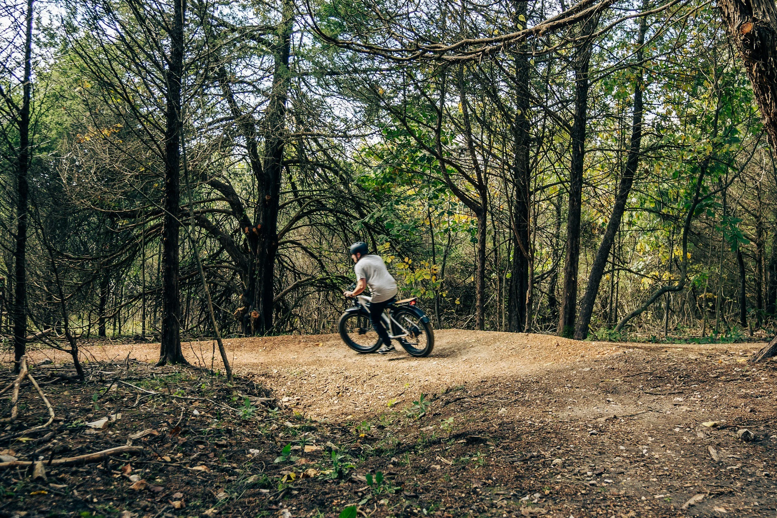 A person riding a mountain bike on a dirt trail through a wooded area with trees and green foliage.