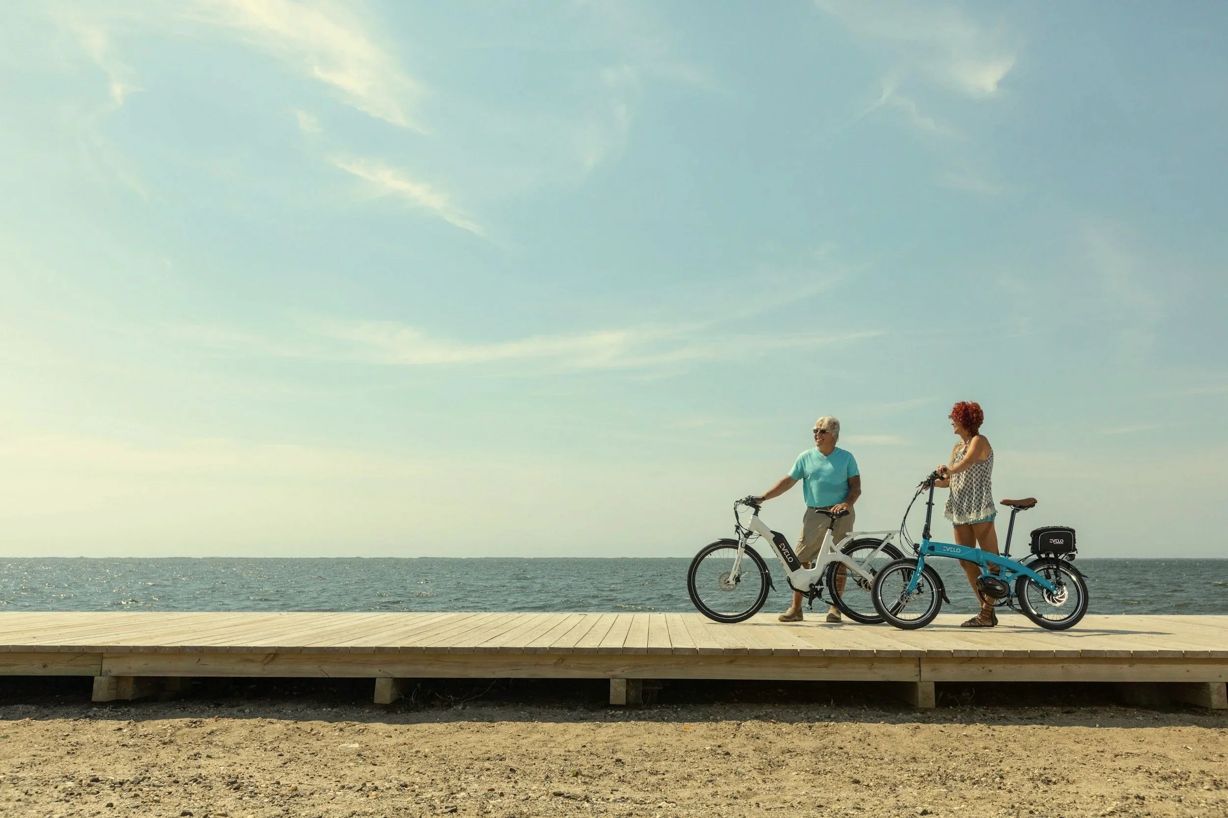 Two women with bicycles standing on a wooden boardwalk by the ocean under a partly cloudy sky.