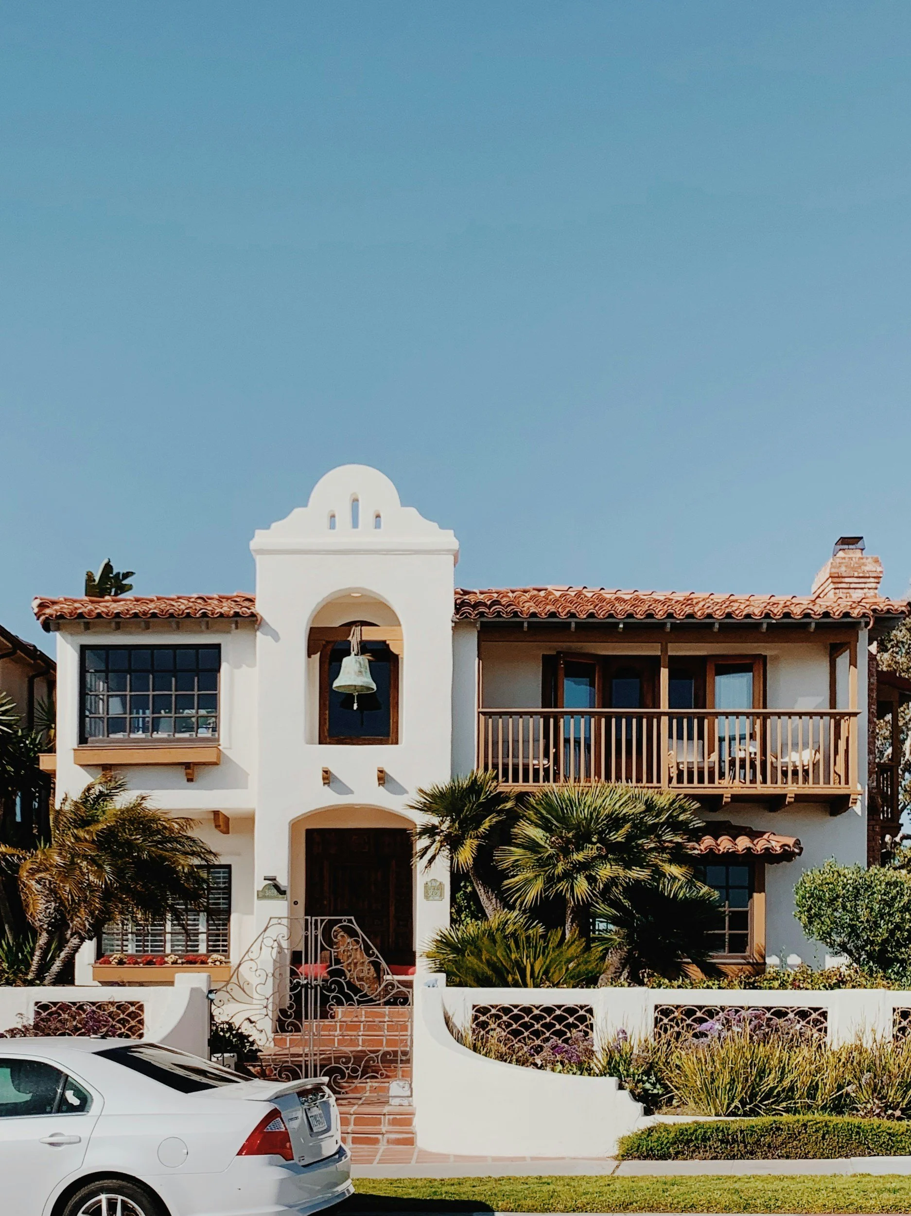 A two-story Mediterranean-style house with a white stucco exterior, red-tile roof, and wooden balcony, surrounded by tropical plants and a white fence, with a car parked in front and a clear blue sky above.