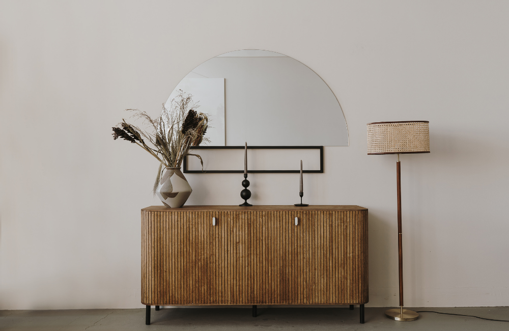 Decorative interior with a wooden sideboard, a geometric vase with dried flowers, two black candles, a half-moon mirror, and a tall floor lamp with a woven shade against a plain white wall.