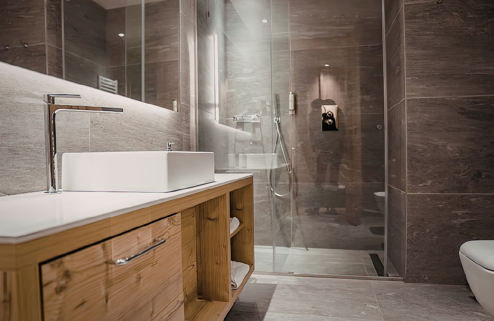 Modern bathroom with a wooden vanity, rectangular vessel sink, and a glass-enclosed shower with dark tile walls.