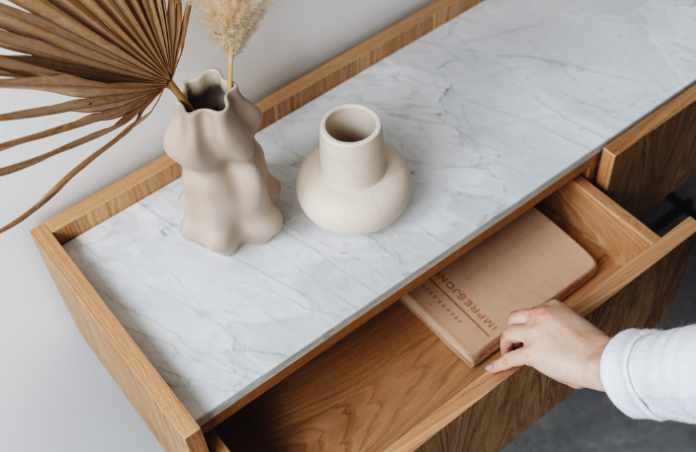 A wooden console table with a white marble top decorated with two ceramic vases and a dried palm leaf. A hand is pulling out a drawer revealing a brown paper package.