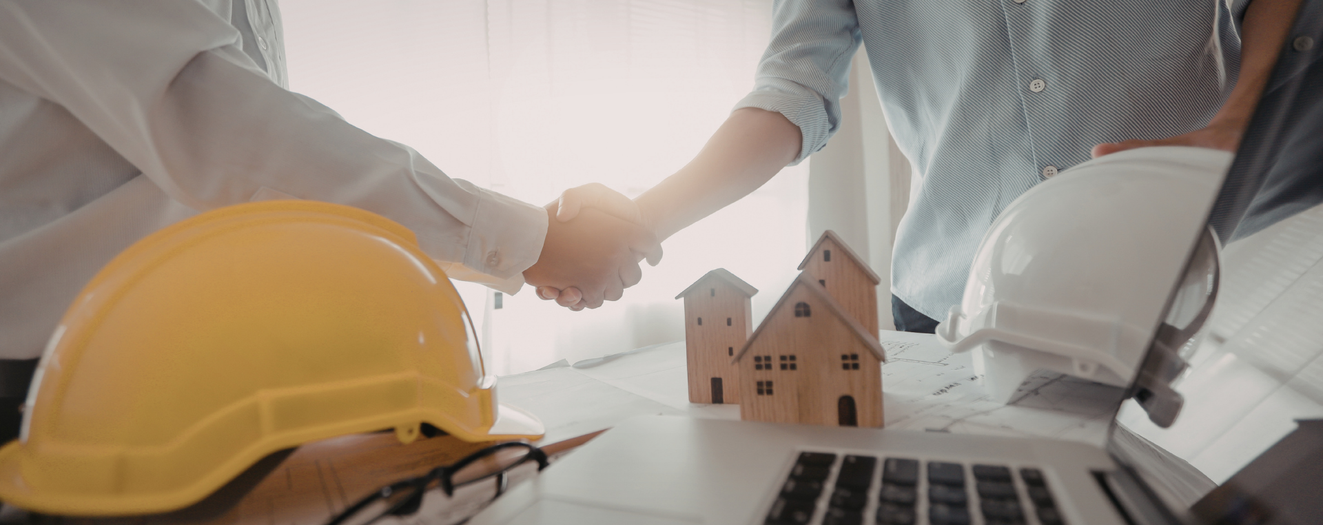 Two people shaking hands over a table with small wooden house models, a yellow construction helmet, a white safety helmet, blueprints, and a laptop.