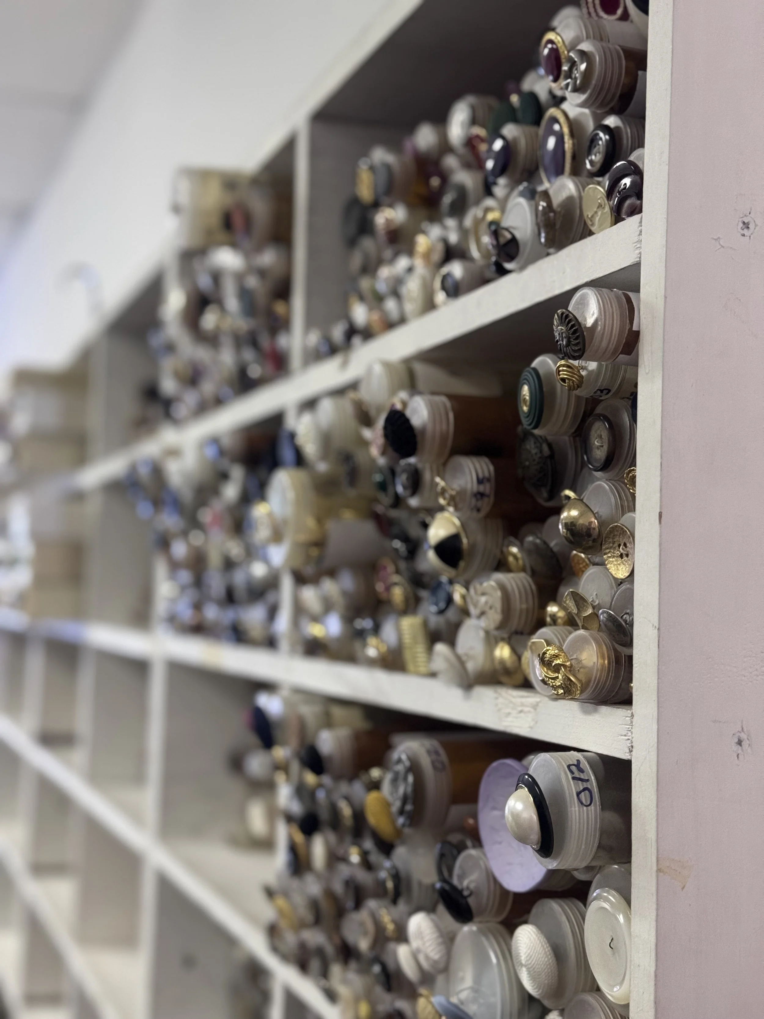 Floor to ceiling white sleving that holds hundreds of tubes of vintage buttons. Each tube has an example button glued to the end facing off the shelf for identification.
