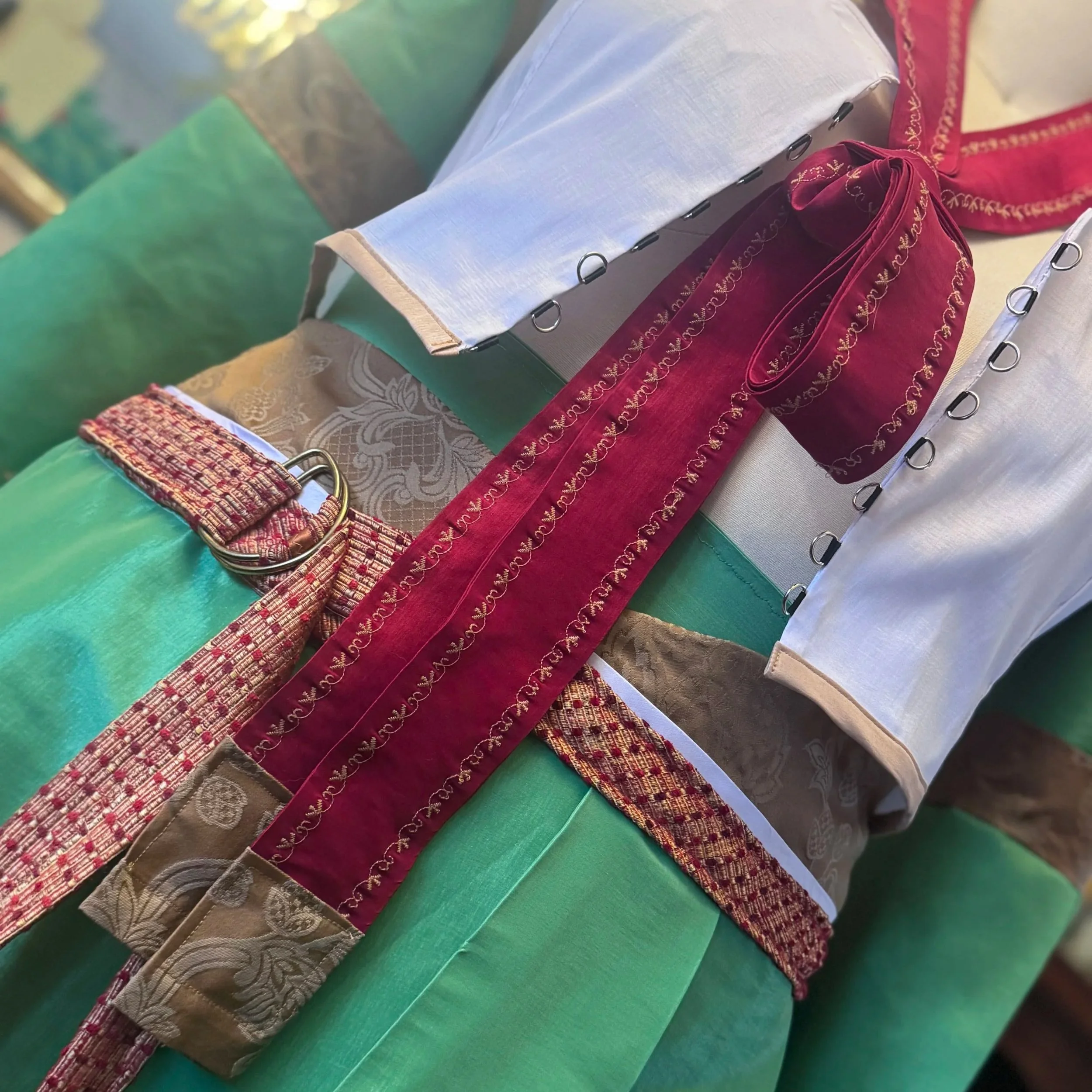 Detail view of scarlet red accessory sash with gold brocade and tassel details as well as gold embroidery.