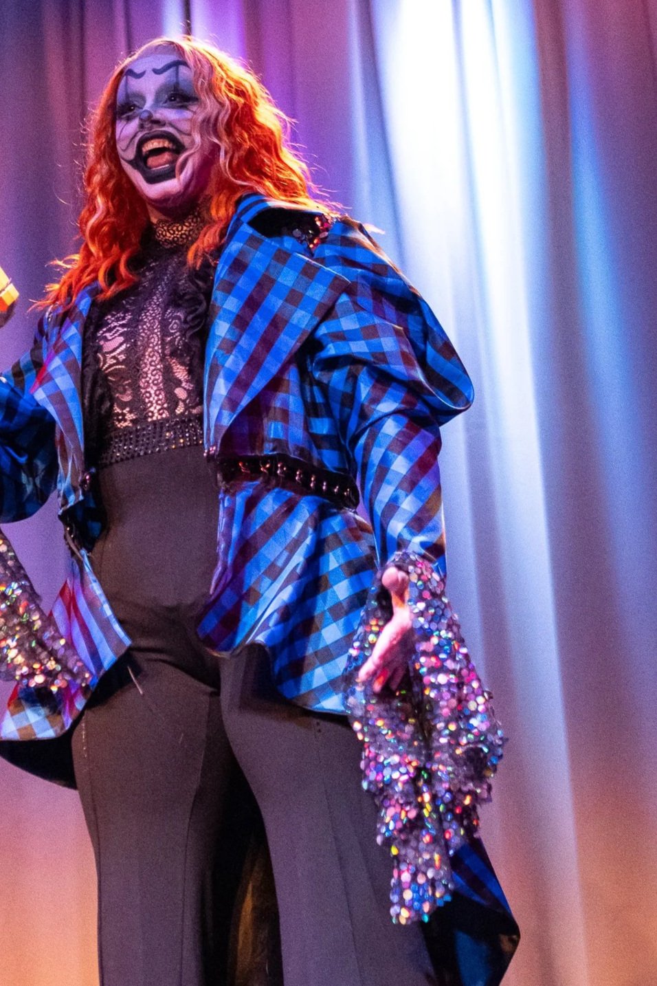 A drag performer with clown makeup and curly red hair on stage, wearing a blue plaid jacket, a lace top, black pants, and holding a sparkly coat.