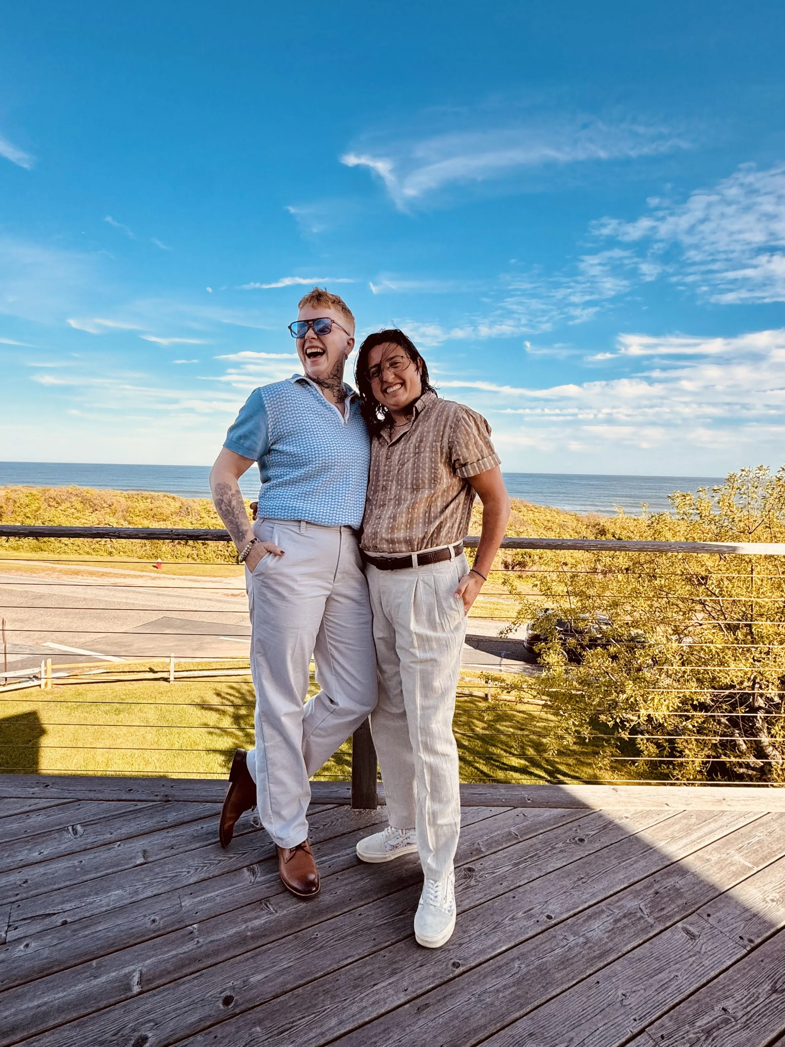 Two people smiling and posing together on a wooden deck with a scenic beach and ocean view under a partly cloudy sky.