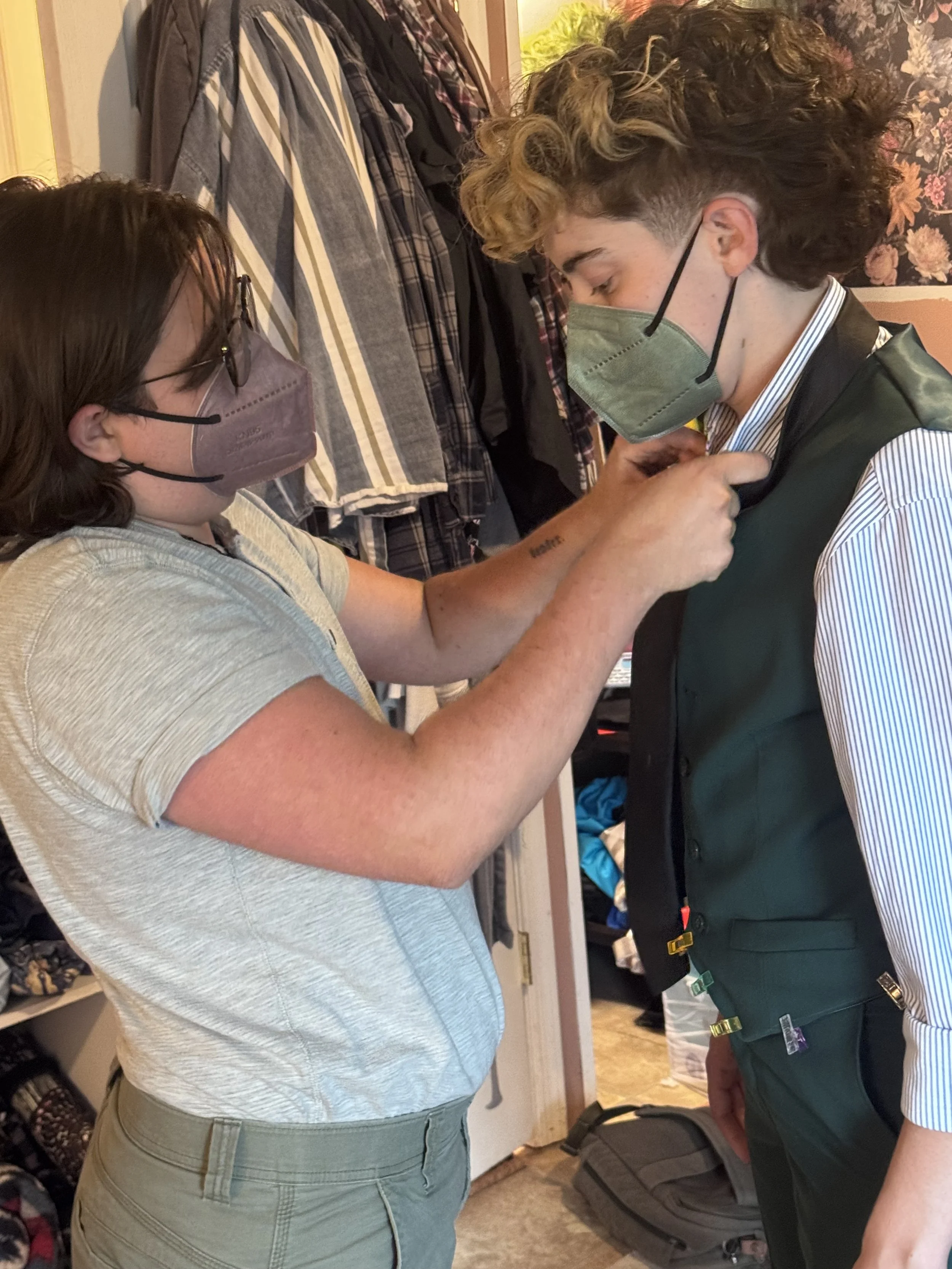 A nonbinary person helping another nonbinary person adjust their clothing and tie their tie, both wearing face masks, in a room with jackets hanging on hooks and shelves in the background.
