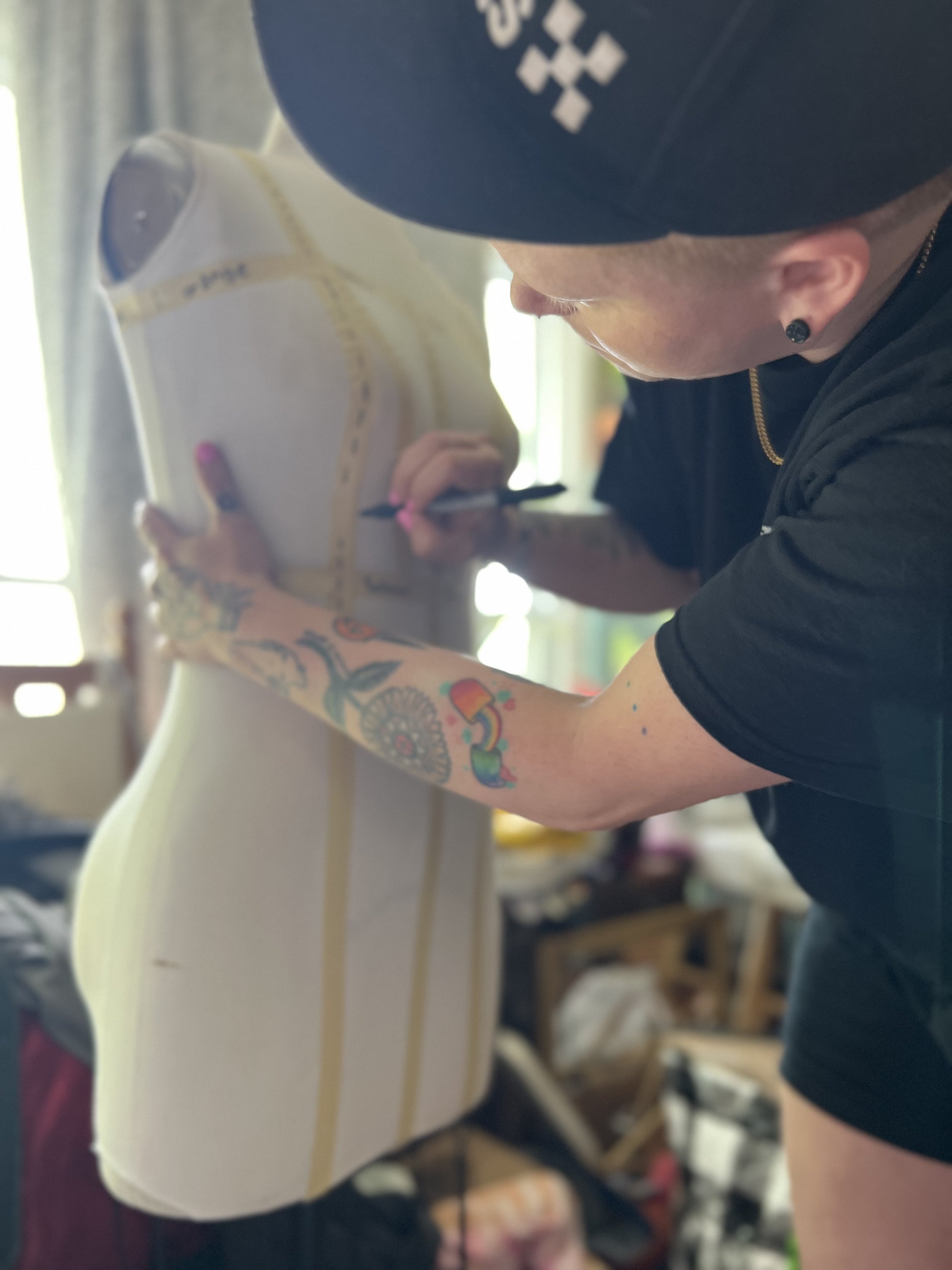 white androgynous person with arm tattoos and black tee shirt wearing a black flatbrim hat and making markings on a size 10 dress form