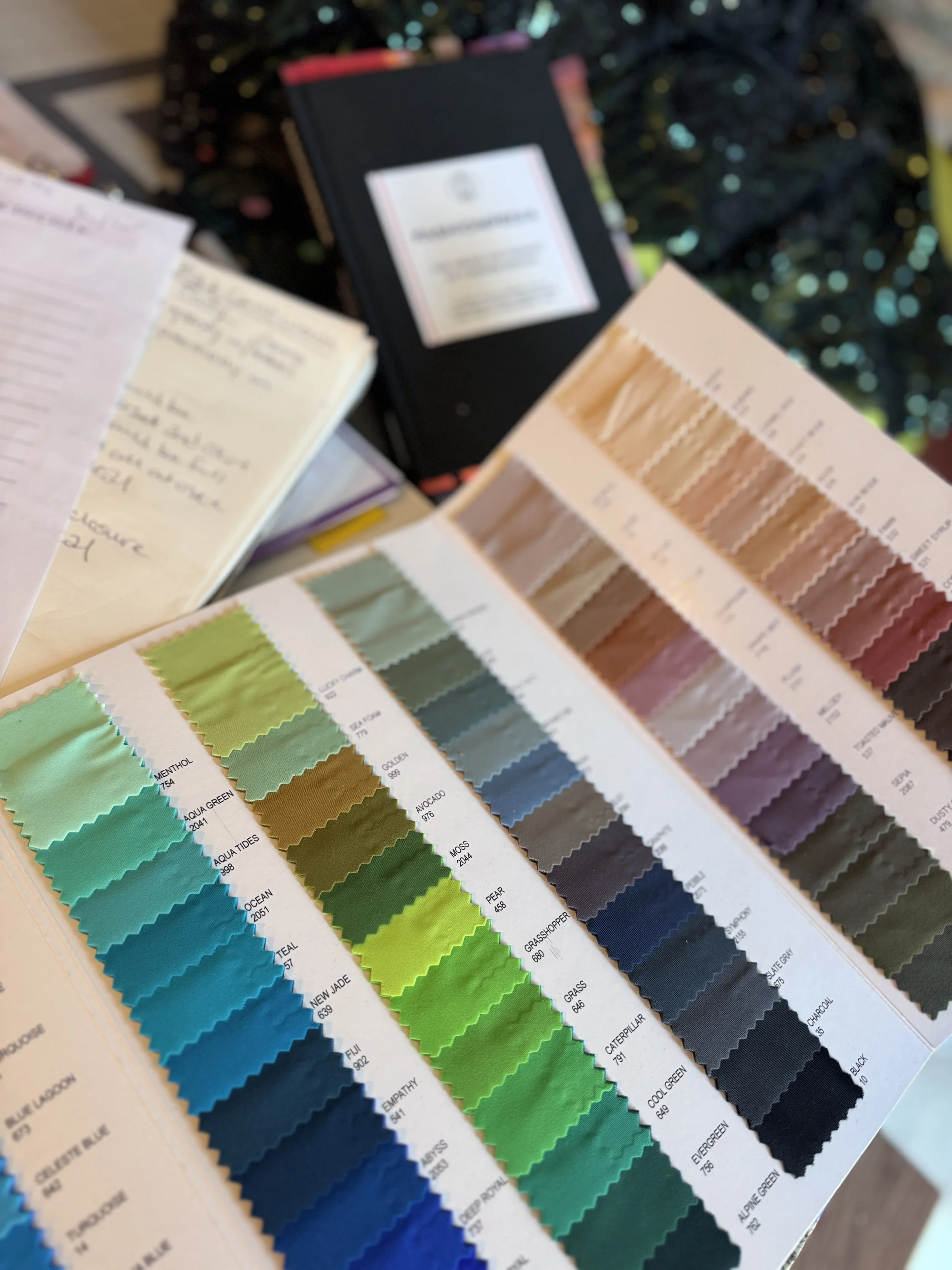 Close-up view of a color palette book showing a range of fabric or paint swatches in shades of green, blue, and earth tones. The palette is displayed with color names and codes.