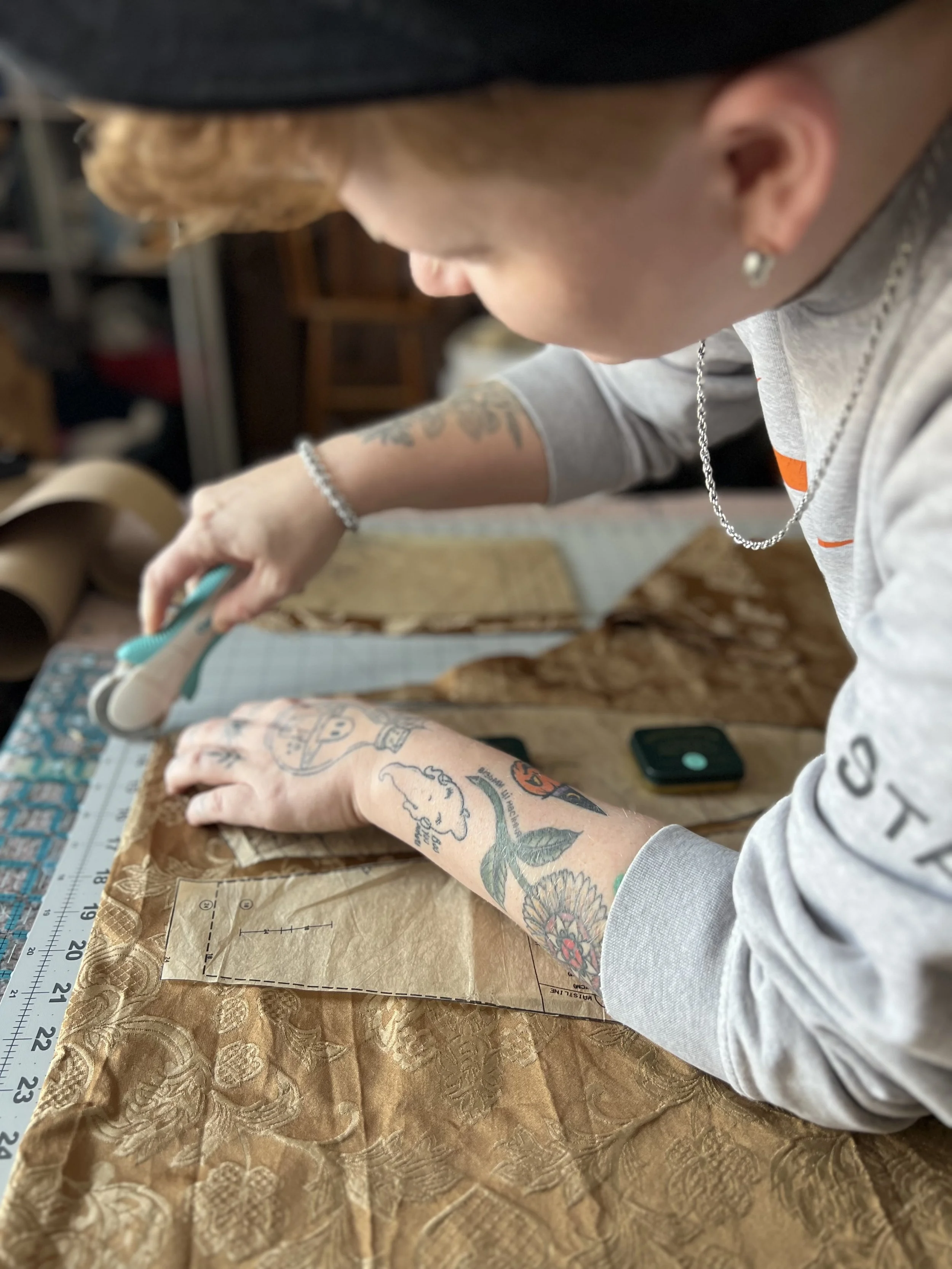 A nonbinary person with short, red hair and tattoos on their arms working on drafting a sewing pattern, using a ruler and fabric with a textured pattern on a work surface.