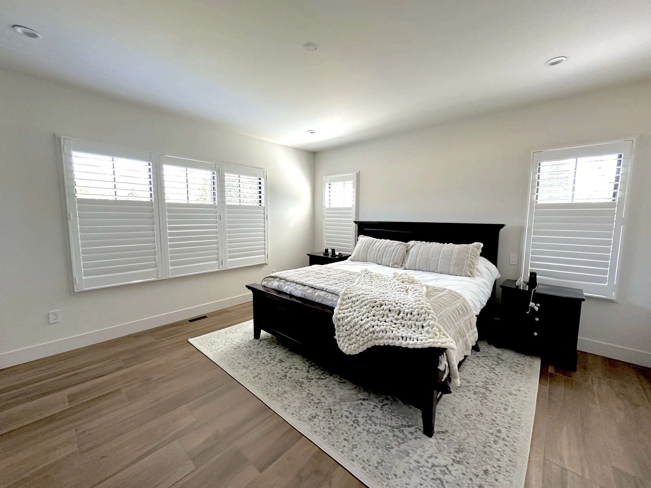 A bedroom with a black bed frame, beige bedding, and a chunky knit blanket. There are black nightstands on each side of the bed, and white plantation shutters on the windows. The room has light-colored walls, hardwood floors, and a light-colored area
