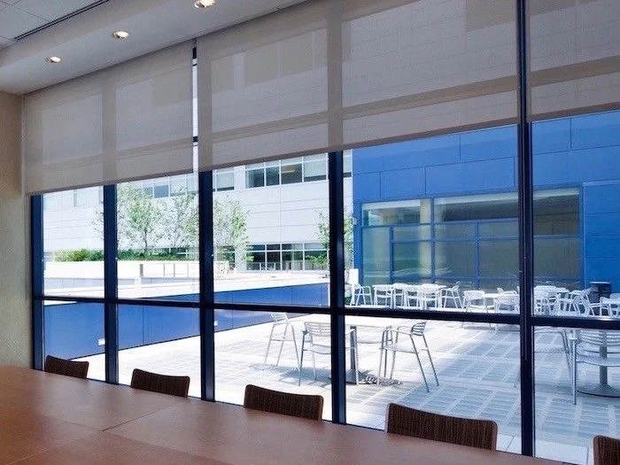 Inside an office conference room with a long wooden table and chairs, view through large windows shows an outdoor patio with tables and chairs, surrounded by modern buildings.