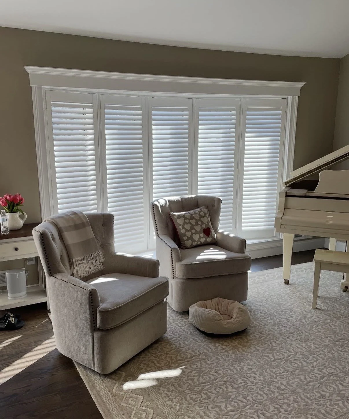Living room with two beige armchairs, a white grand piano, a side table with pink flowers, and a rug, illuminated by sunlight through white plantation shutters.