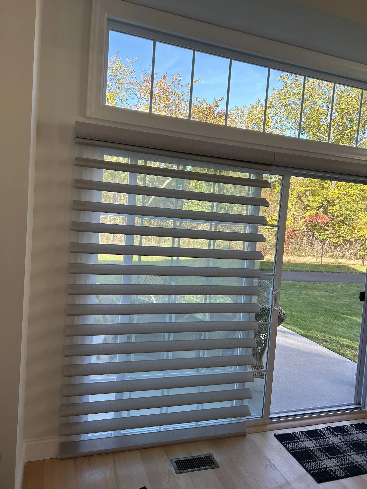 slide patio door with zebra blinds, outside view of trees and grass, wicker chair outside, hardwood floor, and a black and white patterned doormat.