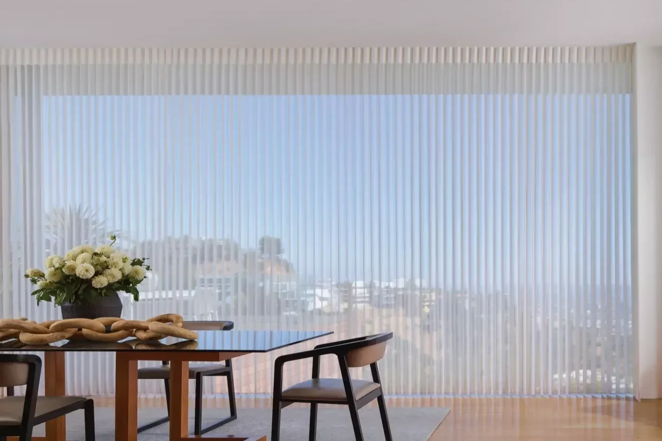 Modern dining room with a wooden table, black chairs, a floral centerpiece, and floor-to-ceiling vertical blinds covering large windows with a city view.