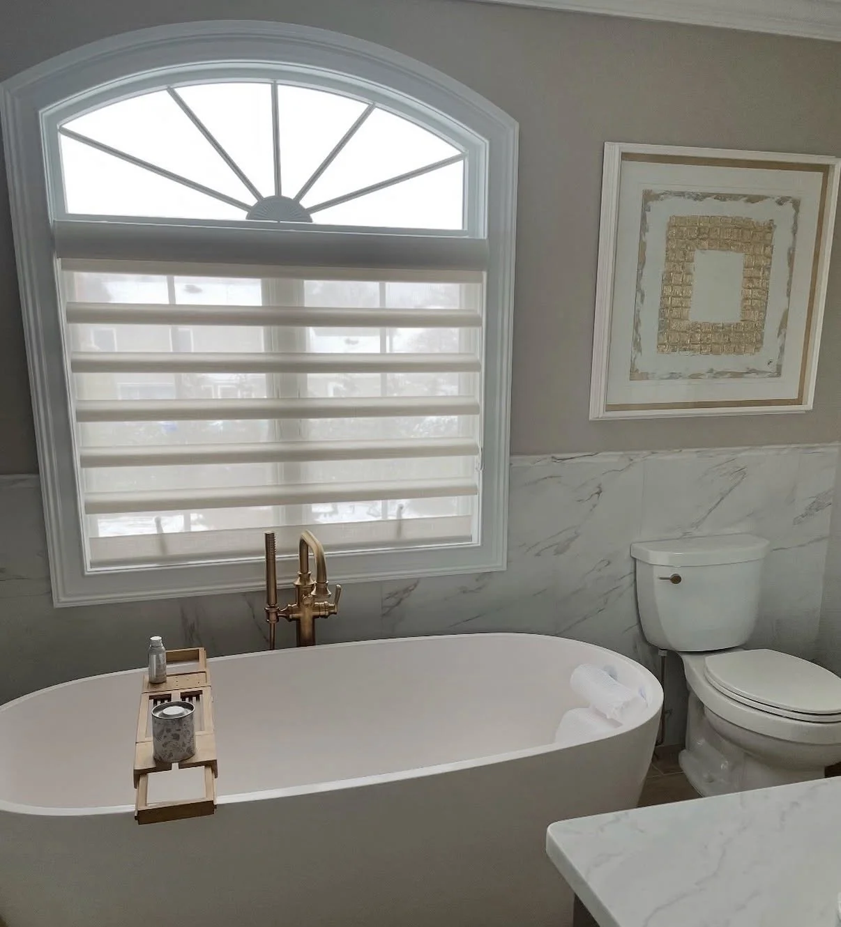 Bathroom featuring a freestanding bathtub with a gold faucet near a large window with striped shades. There is a toilet next to the bathtub and a framed artwork on the wall. The room has marble wall accents.