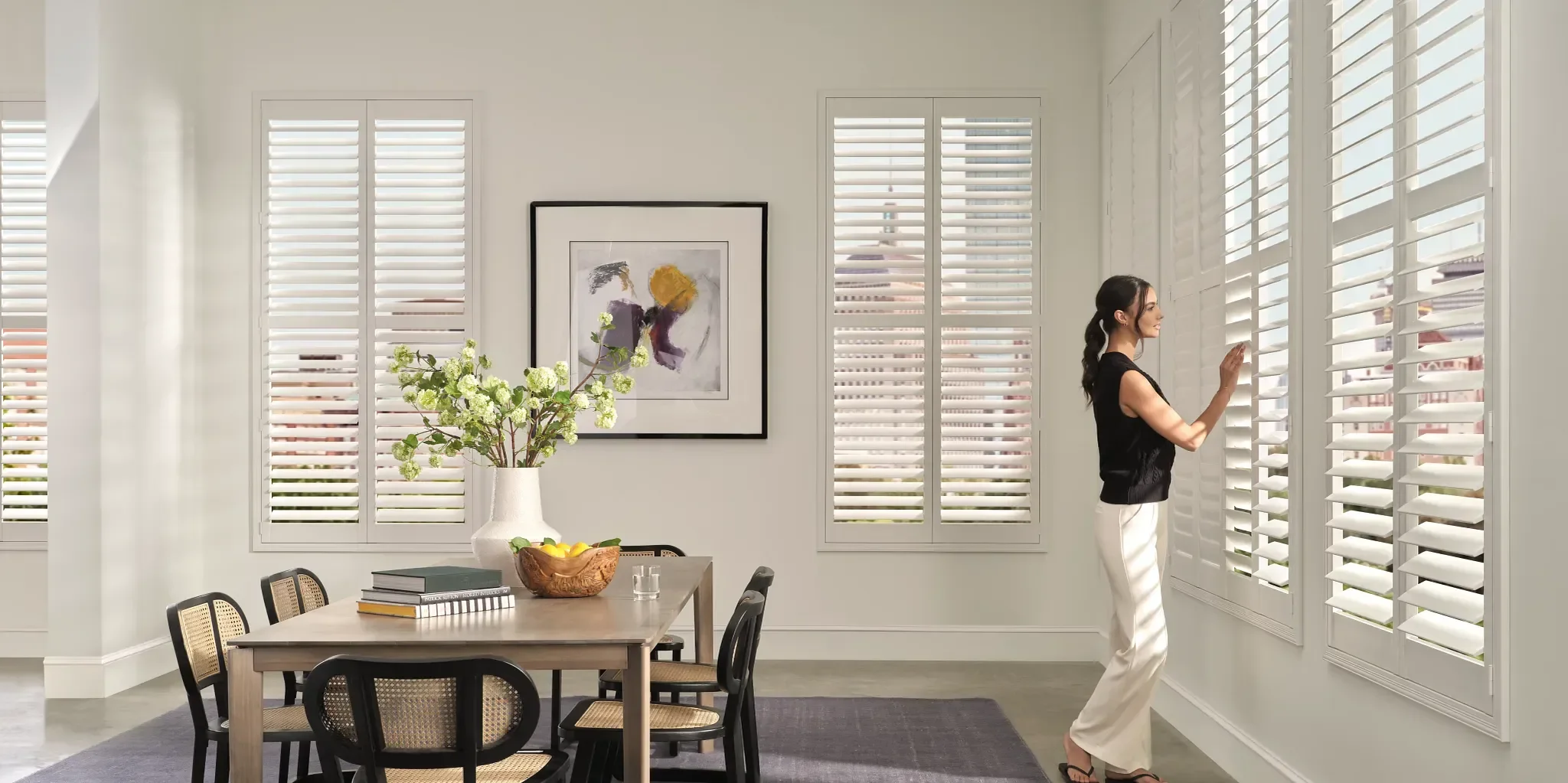 Woman standing near window with white plantation shutters in dining room, with a wooden table, chairs, a large vase with white flowers, and framed artwork on the wall.