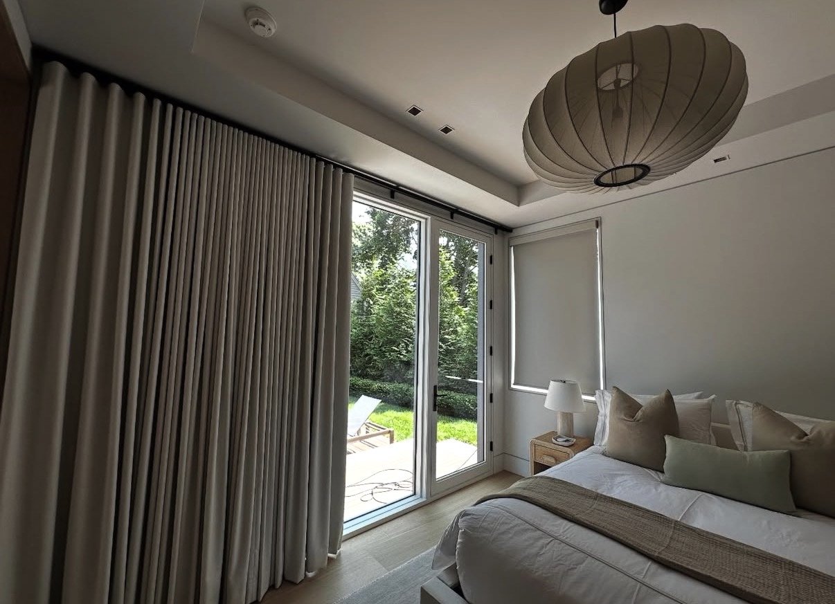 A bedroom with a large bed, pillows, a nightstand with a lamp, and a ceiling light fixture. There's a sliding glass door with curtains and a window with shades, revealing a backyard with trees and outdoor furniture.