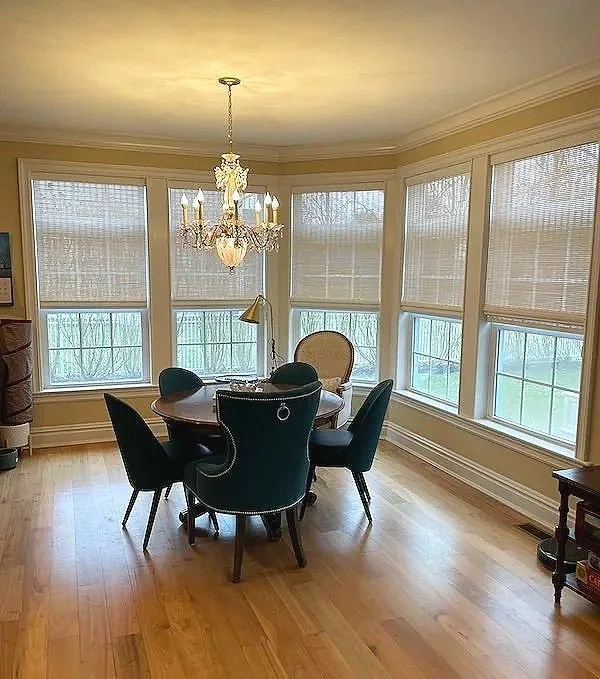 Elegant dining room with round table, six chairs, chandelier, hardwood floor, large windows with blinds, and a small side table.