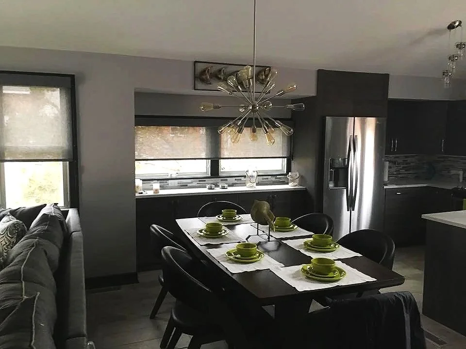Modern kitchen and dining area with a black dining table, six green bowls and plates, a silver globe-shaped chandelier, large windows with blinds, and gray and black cabinets, refrigerator, and sofa.