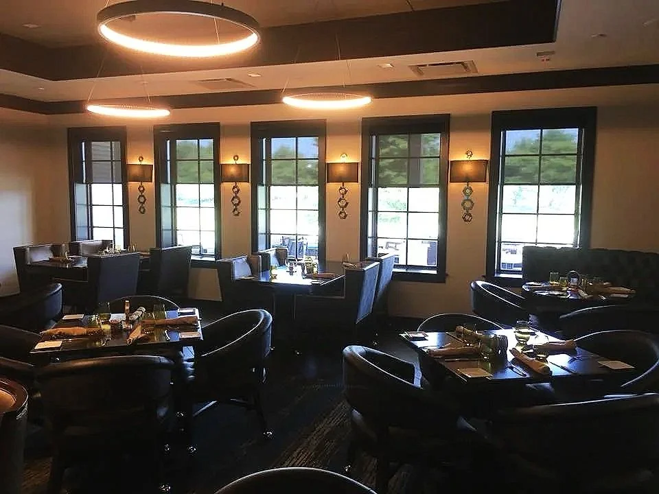 Interior of a restaurant with multiple tables set with plates, glasses, and silverware. Leather chairs surround the tables, and large windows let in natural light. Modern ceiling lights brighten the space.