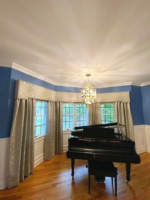 A black grand piano in a bright room with blue walls, white trim, and large windows with patterned curtains, illuminated by a decorative chandelier.