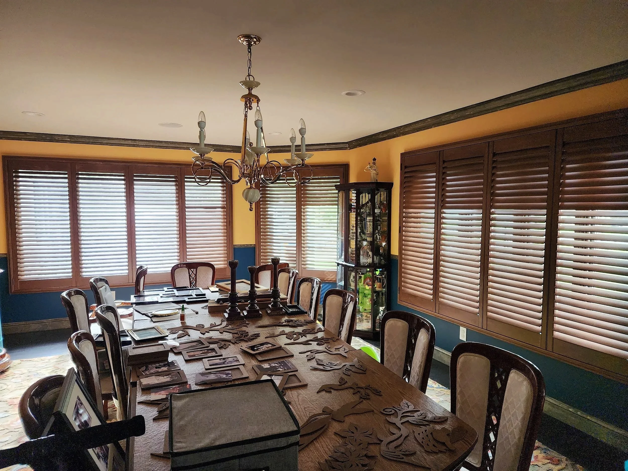 A dining room with a large wooden table surrounded by upholstered chairs, a chandelier hanging overhead, and multiple windows with wooden blinds. Decorative items on the table and a glass cabinet in the corner.