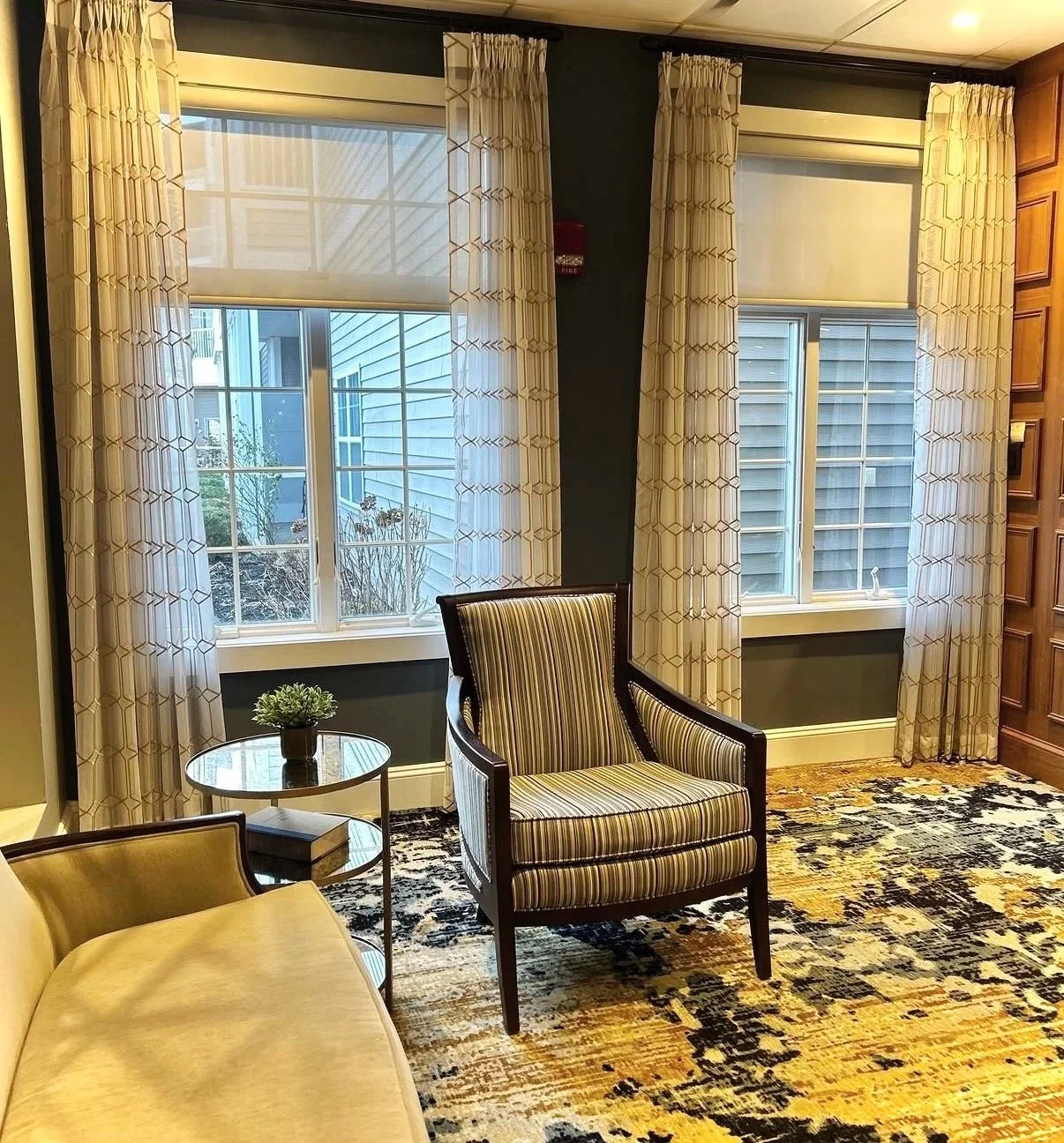 Interior of a living room with two large windows, beige curtains, a striped upholstered armchair, a side table with a plant, a beige sofa, and a patterned area rug.