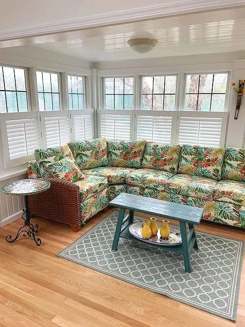 Sunroom with large tropical print sofa, window shutters, a small side table, and a coffee table with yellow vases, on a patterned rug.