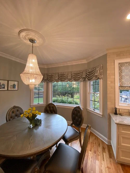 A dining area with a chandelier, a wooden table, and four chairs, one with a tufted backrest, decorated with a yellow flower bouquet. There are three windows with draped valances showing a green outdoor view.