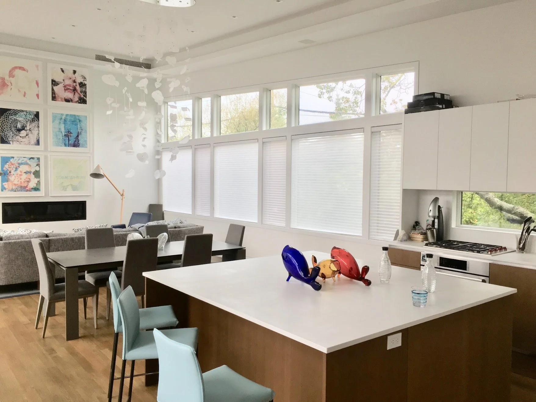 Modern open-concept kitchen and living room with a white island, light blue barstools, a dark dining table with six gray chairs, and a gray sofa. Wall art is displayed on a gallery wall, and a white ceiling installation with hanging paper shapes. Lar