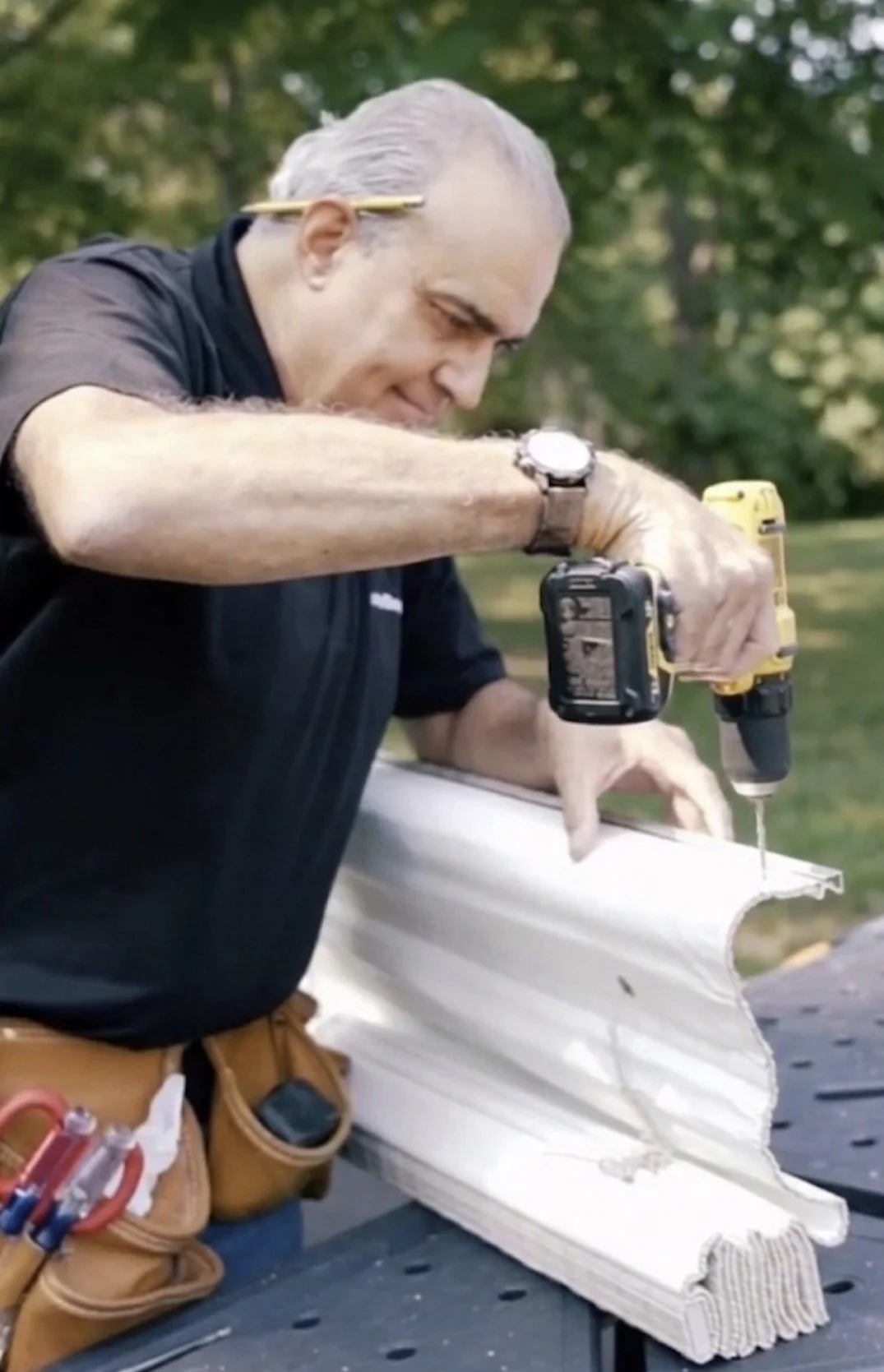 Man working on a white architectural model with a power drill outdoors, wearing a tool belt and watch.