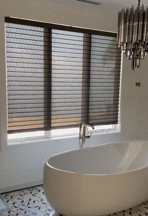 Modern bathroom with a freestanding white bathtub, large window with gray horizontal blinds, and a geometric black and white tiled floor. There is a chrome faucet and a metallic chandelier hanging from the ceiling.